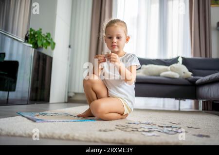 Das kleine blonde Mädchen sitzt zu Hause auf dem Teppich und sammelt Rätsel Stockfoto