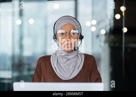 Junge muslimische Frau im Hijab, die geschäftliche Schulungen und Online-Webinare im Büro durchführt und ernsthaft am Tisch im Headset vor dem Laptop sitzt. Nahaufnahme. Stockfoto