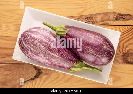 Zwei reife Auberginen mit weißer Keramikplatte auf Holztisch, Makro, Draufsicht. Stockfoto