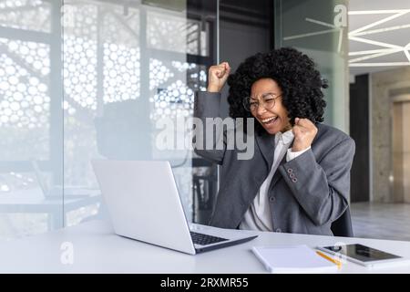 Erfolgreiche afroamerikanische Frau am Arbeitsplatz innerhalb des Büros, Geschäftsfrau Gewinnerin erhielt gute Ergebnisse und Leistung am Arbeitsplatz, indem sie Benachrichtigung mit Laptop las. Stockfoto