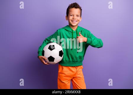 Foto eines fröhlichen, selbstbewussten, grünen Hoodies für kleine Jungen, der den Daumen hoch hält und einen isolierten violetten Hintergrund zeigt Stockfoto