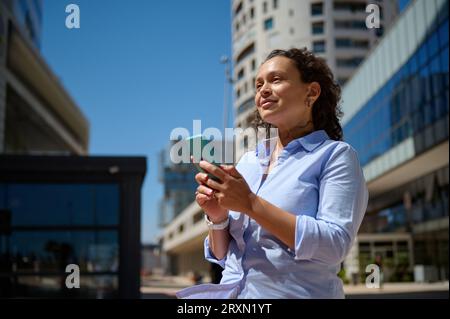 Schöne Managerin in Freizeitkleidung, Unternehmerin, Freiberuflerin, Geschäftsfrau mit Mobiltelefon, lächelnd und selbstbewusst beiseite schauend, stehend A Stockfoto