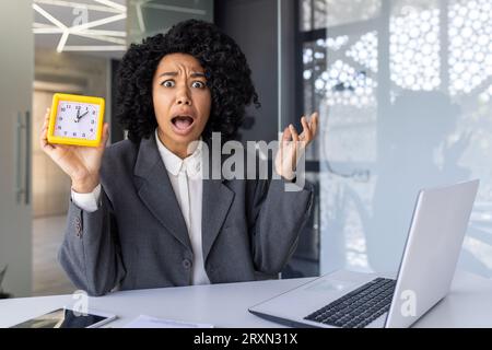 Überarbeitete Frau, die spät arbeitet, Geschäftsfrau, die auf die Kamera schaut und die Uhr zeigt, Arbeiterin mit Laptop mitten im Büro müde. Stockfoto