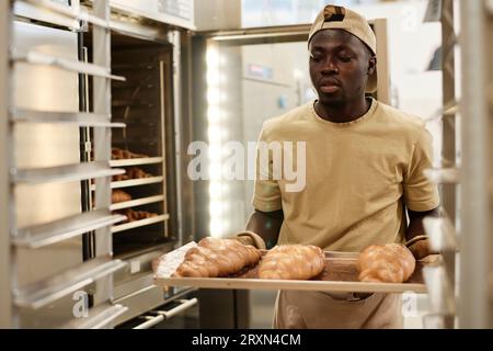 Porträt des schwarzen jungen Mannes, der ein Tablett mit frisch gebackenem Brot in der Backküche hält, Kopierraum Stockfoto