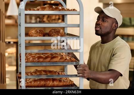 Porträt des jungen Mannes, der in der Backküche arbeitet und das Regal mit frischen Broten und Kopierraum bewegt Stockfoto