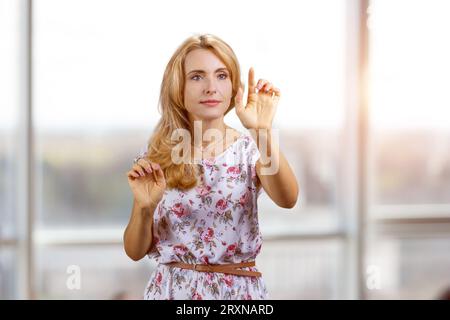 Blonde Frau mittleren Alters mit einem unsichtbaren virtuellen Bildschirm in Innenräumen. Platz für einen Kopierbereich. Verschwommenes Fenster im Hintergrund. Stockfoto