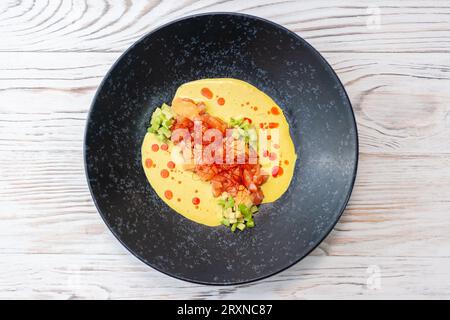 Lachstartar mit Avocado, viel Soße und Chilifäden auf einem schwarzen Teller auf hellem Holzgrund Stockfoto