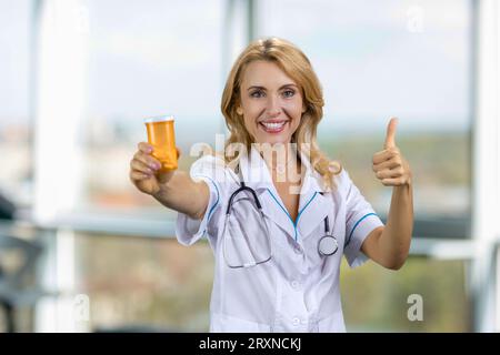 Glücklicher blonder kaukasischer Arzt gibt Ihnen eine Medizinflasche und Daumen hoch. Verschwommenes Fenster im Hintergrund. Stockfoto