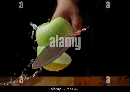 Frau schneidet einen Apfel in zwei Hälften mit einem Messer in der Luft, das Wasser verschüttet, Spritzeffekt, isoliert auf schwarzem Hintergrund. Hochgeschwindigkeitsfotografie Stockfoto