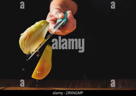 Frau schneidet eine Birne in zwei Hälften mit einem Messer in der Luft, das Wasser verschüttet, Spritzeffekt, isoliert auf schwarzem Hintergrund. Hochgeschwindigkeitsfotografie Stockfoto