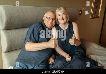 Ein älteres Paar sitzt auf einem Bett und zeigt eine Geste mit den Händen - Daumen hoch, wie. Stockfoto