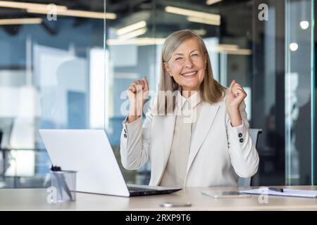 Porträt einer erfolgreichen Geschäftsfrau mit grauen Haaren, weiblicher Boss lächelnd und mit Blick auf die Kamera, Hände hochhaltend, Gewinnergeste, zufrieden mit den Erfolgen, triumphierend. Stockfoto