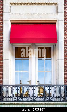 Hohe Doppeltüren mit Glas und einem hellroten textilen Baldachin öffnen sich auf einen kleinen Balkon mit schmiedeeisernen Metallgeländern in einem klassischen Backsteingebäude Stockfoto