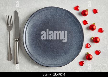 Leere blaue Platte mit Besteck, roten Weihnachtskugeln und Herzen auf einem grauen Steintisch. Festliches Neujahrsmenü. Stockfoto