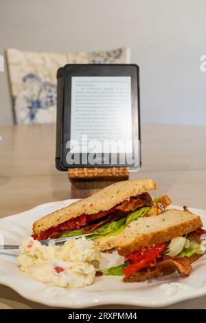 Eine Mahlzeit, während Sie ein Buch über ein Kindle lesen, ein BLT essen, ein Sandwich mit Speck, Tomaten und Salat, das mit Getreidebrot zubereitet und auf einem weißen Teller serviert wird. USA. Stockfoto