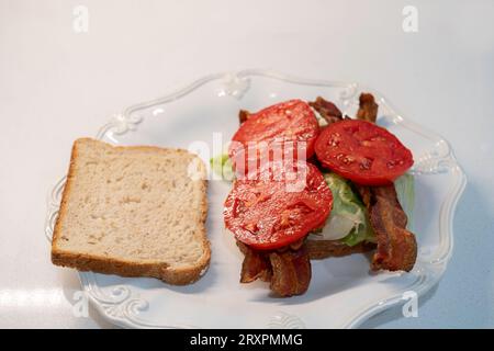 BLT, ein Sandwich mit Speck, Tomaten und Salat, das mit Haferbrot zubereitet wird, wobei eine Brotscheibe entfernt wird, die den Inhalt zeigt, auf einem weißen Teller. USA. Stockfoto