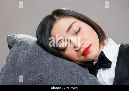 Müde, verschlafene Frau an der Rezeption mit geschlossenen Augen, die kopfüber auf dem Kissen liegen. Restaurant-Catering-Service junge, attraktive, erschöpfte asiatische Kellnerin schläft und entspannt in der Nähe Stockfoto