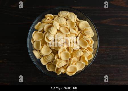 Getrocknete gelbe Orecchiette-Pasta in einer Glasschale: Eine Schüssel ungekochter Orecchiette-Pasta auf einem Holztisch von oben gesehen Stockfoto