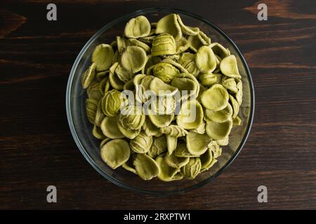 Getrocknete grüne Orecchiette-Pasta in einer Glasschale: Eine Schüssel ungekochter Orecchiette-Pasta auf einem Holztisch, von oben gesehen Stockfoto