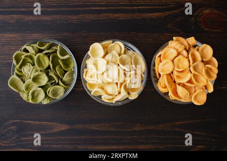 Getrocknete dreifarbige Orecchiette-Pasta in einer Glasschale: Drei Schalen ungekochter Orecchiette-Pasta auf einem Holztisch, von oben gesehen Stockfoto