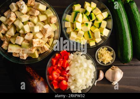 Gehacktes Sommergemüse in Glasschüsseln: Vorbereitete Aubergine, Zucchini, rote Paprika, Zwiebeln und Knoblauch auf einem Holztisch Stockfoto