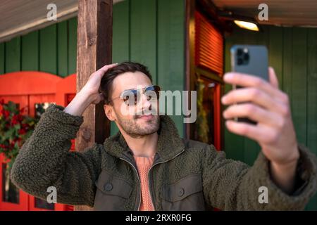 Mann, der im Freien Selfie macht. Stilvoller Mann, der eine Sonnenbrille trägt und mit dem Handy vor dem Holzhaus ein Foto von sich macht. Seitenansicht eines hübschen Mannes in einer flauschigen Jacke, der draußen Videochat führt Stockfoto
