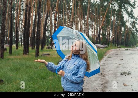 Junge Frau, die einen durchsichtigen blauen Regenschirm im Wald hält. Regnerischer Tag mit Regenschirm. Frau mit der Hand, die prüft, wie lange es regnet. Prognose Stockfoto