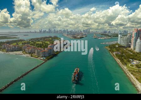 Kommerzielles Containerschiff, das den Hafen von Miami über den Hauptkanal in der Nähe von South Beach erreicht. Luxuriöse Hotels und Wohngebäude am Wasser Stockfoto