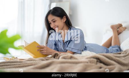 Eine attraktive und glückliche asiatische Frau im Pyjama liest gerne morgens ein Buch auf ihrem Bett im Schlafzimmer. Freizeit- und Lifestyle-Konzept Stockfoto