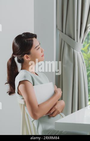 Eine junge romantische Frau, die im Wohnzimmer ein Buch an die Brust hält Stockfoto