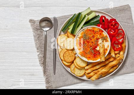 Hot Pepper Jelly Cheese Dip in Bowl serviert mit Crackern, knackigen italienischen Brotstangen, frischem Pfeffer und Gurke auf Platte auf weißem Holztisch, Horiz Stockfoto