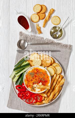 Hot Pepper Jelly Cheese Dip in Bowl serviert mit Crackern, salzigem grissini oder knackigen italienischen Brotstangen, frischem Pfeffer und Gurke auf Platte auf weißem W Stockfoto