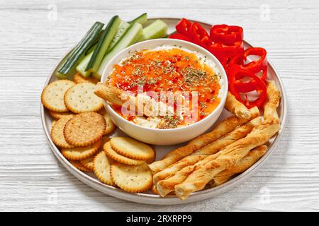 Hot Pepper Jelly Cheese Dip in Bowl serviert mit Crackern, salzigem grissini, frischem Pfeffer und Gurke auf Platte auf weißem Holztisch Stockfoto