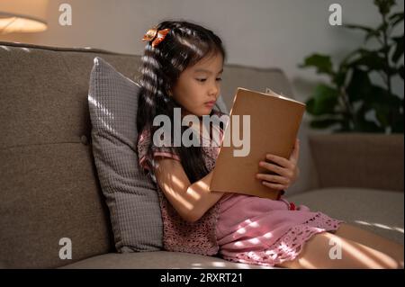 Ein süßes kleines asiatisches Mädchen konzentriert sich darauf, ein Buch oder ein Märchen auf einer Couch im Wohnzimmer zu lesen. Freizeit- und Hobbykonzepte Stockfoto