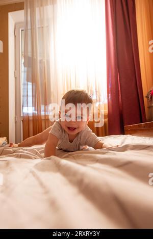 Ein einjähriges Baby liegt morgens in der Sonne auf dem Bauch auf dem Bett. Das Kind ist lustig, lächelt, spielt in guter Stimmung. Pflege, Möbel für Stockfoto