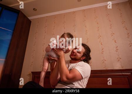 Ein vielseitiger Vater mit langen Haaren und Brille wirft den kleinen Sohn auf das Bett. Ein junger Mann rollt einen Jungen in die Arme, ein Kind gibt vor, ein Flugzeug zu sein Stockfoto