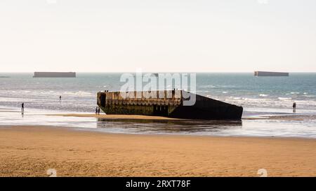 Ein Element des Mulberry-Hafens aus dem Zweiten Weltkrieg, erbaut am Gold Beach in Arromanches, Frankreich, liegt auf dem Sand mit Überresten von Phoenix Caissons in der Ferne. Stockfoto