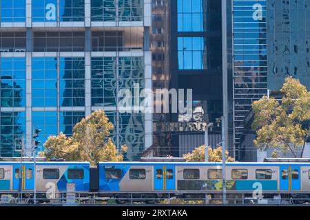 Öffentliche Verkehrsmittel Melbourne zeigt einen elektrischen Personenzug, der auf Bahngleisen mit Glasfenstern und Stadtgebäuden fährt. Stockfoto