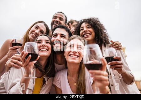 Unterer Blick auf eine multirassische Gruppe von Freunden, die auf einer Dachparty Getränke trinken. Vielfältige junge glückliche Gruppe von Millennials, die Wein auf einer Terrasse trinken Stockfoto