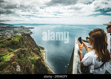 Panoramablick auf ein Paar, das mutig die atemberaubende Aussicht vom Cabo Girão Skywalk, Madeira, Portugal genießt und Bilder von der Landschaft mit dem Handy macht Stockfoto