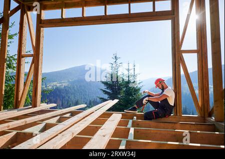 Zimmermann bauend Holzrahmen zweistöckiges Haus in der Nähe des Waldes. Bärtiger Mann mit Hammer, Schutzhelm und Overall. Konzept des ökologischen modernen Bauens. Stockfoto