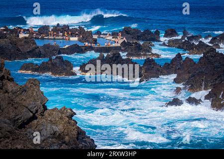 Blick auf viele Menschen, die am Rand eines natürlichen Lavapads in Porto Moniz, Madeira, Portugal stehen, während das Meer um sie herum tobt Stockfoto