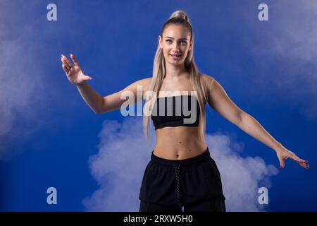 Schönes blondes Mädchen in schwarzem Outfit, das vor Rauchnebel und blauem Hintergrund tanzt. Stockfoto