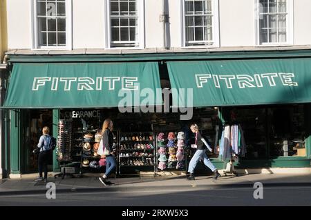 Junge Frauen, die an der Vorderseite des Fitrite-Geschäfts vorbeilaufen, mit einer farbenfrohen Auswahl an Schuhen, Hüten, Kleidung und Sonnenbrillen draußen. Oxford, England, Großbritannien Stockfoto