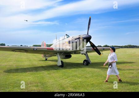 Eine Frau in alten Gewändern, die an einem Hawker Hurricane Mk.1 P2921 GZ-L vorbeiläuft, das auf Gras bei Goodwood Revival, West Sussex, England, Großbritannien, geparkt ist Stockfoto