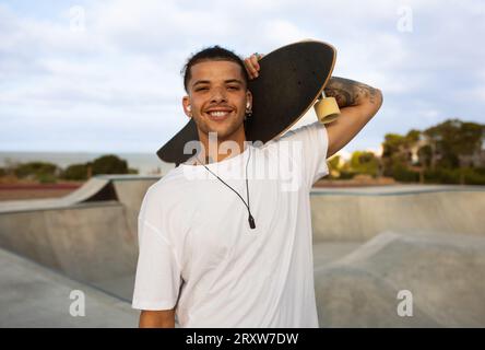 Porträt eines glücklichen, mit Hipster tätowierten europäischen Mannes in einem weißen T-Shirt, das mit Skateboard posiert und im Skatepark spaziert Stockfoto