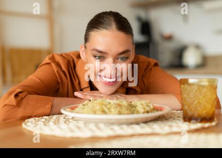 Eine fröhliche junge Frau, die am Tisch sitzt und leckere Pasta auf dem Teller sieht, will Spaghetti essen, Kücheneinrichtung Stockfoto