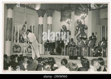 Foto von Cab Calloway auf der Bühne vor seiner Band. Calloway trägt einen hellen Anzug und wird in ein Mikrofon singend dargestellt. Eine große Gruppe sichtbarer Personen, die sitzend auf der Bühne sitzen, entblößt die Bühne. Bandmitglieder auf der Bühne tragen passende dunkle Suiten mit Schleifen. Stockfoto