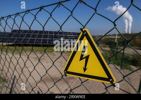 27. September 2023, Sachsen, Groitzsch: Ein Warnschild warnt vor Hochspannungsstrom in der neuen Photovoltaikanlage Peres II im ehemaligen Tagebau vor dem Hintergrund des Braunkohlekraftwerks Lippendorf. Das Bergbauunternehmen Mibrag hat auf einem alten Tagebau südlich von Leipzig eine riesige Photovoltaikanlage in Betrieb genommen. Mehr als 66.000 Solarmodule stehen laut dem Unternehmen auf einer Fläche von 55 Hektar. Das neue Werk Peres II kann jährlich rund 38.000 Megawattstunden Strom produzieren. Vorerst wird Mibrag den Strom intern nutzen, später wird grüner Wasserstoff pro sein Stockfoto