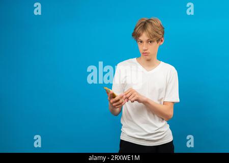 Junge Teenager, die ihr Handy vor blauem Hintergrund benutzen, ihr Mann schreibt online, scrollt Nachrichten, surft auf dem Handy im Internet, genießt das Chatten Stockfoto